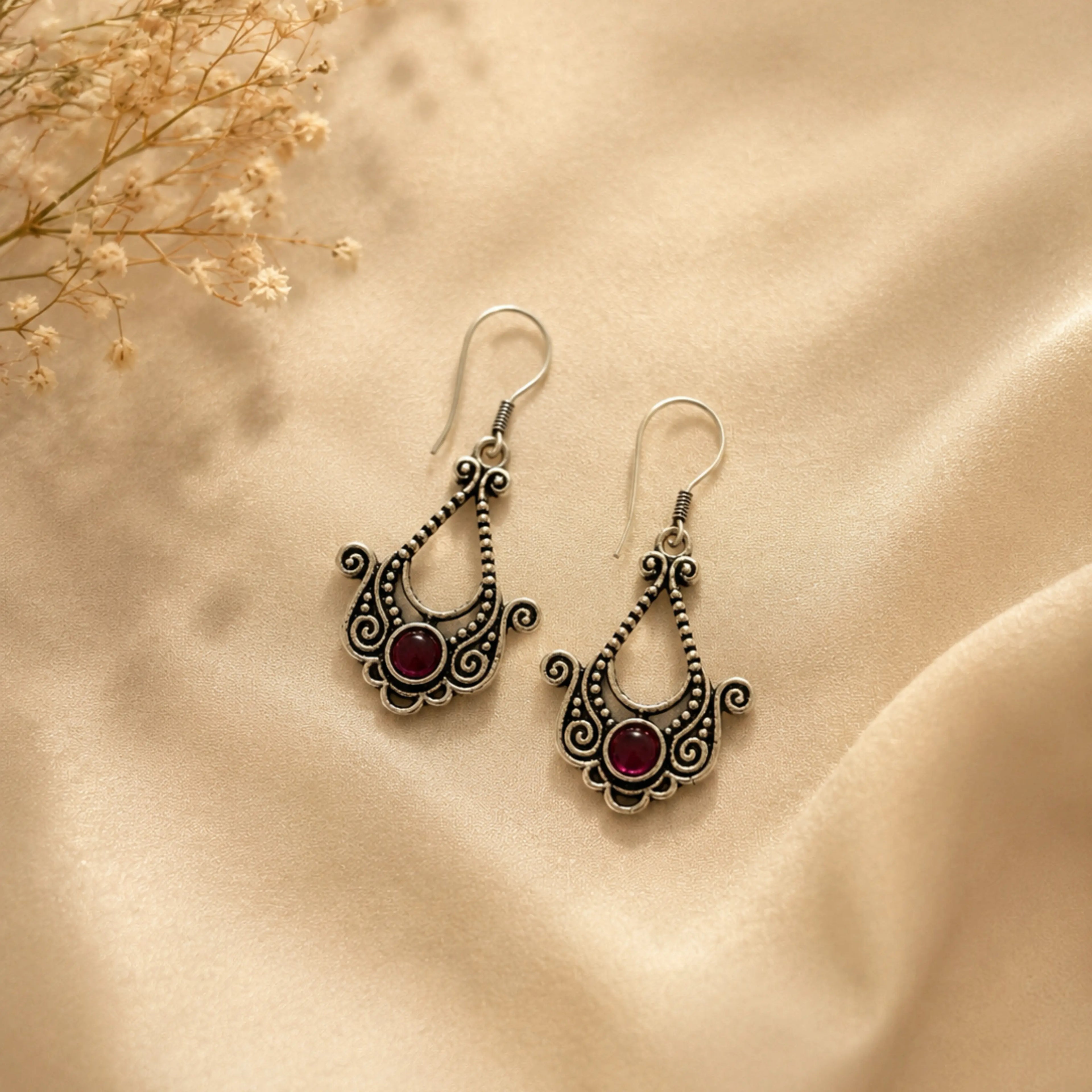 Ornate silver earrings with red stones on a beige fabric background