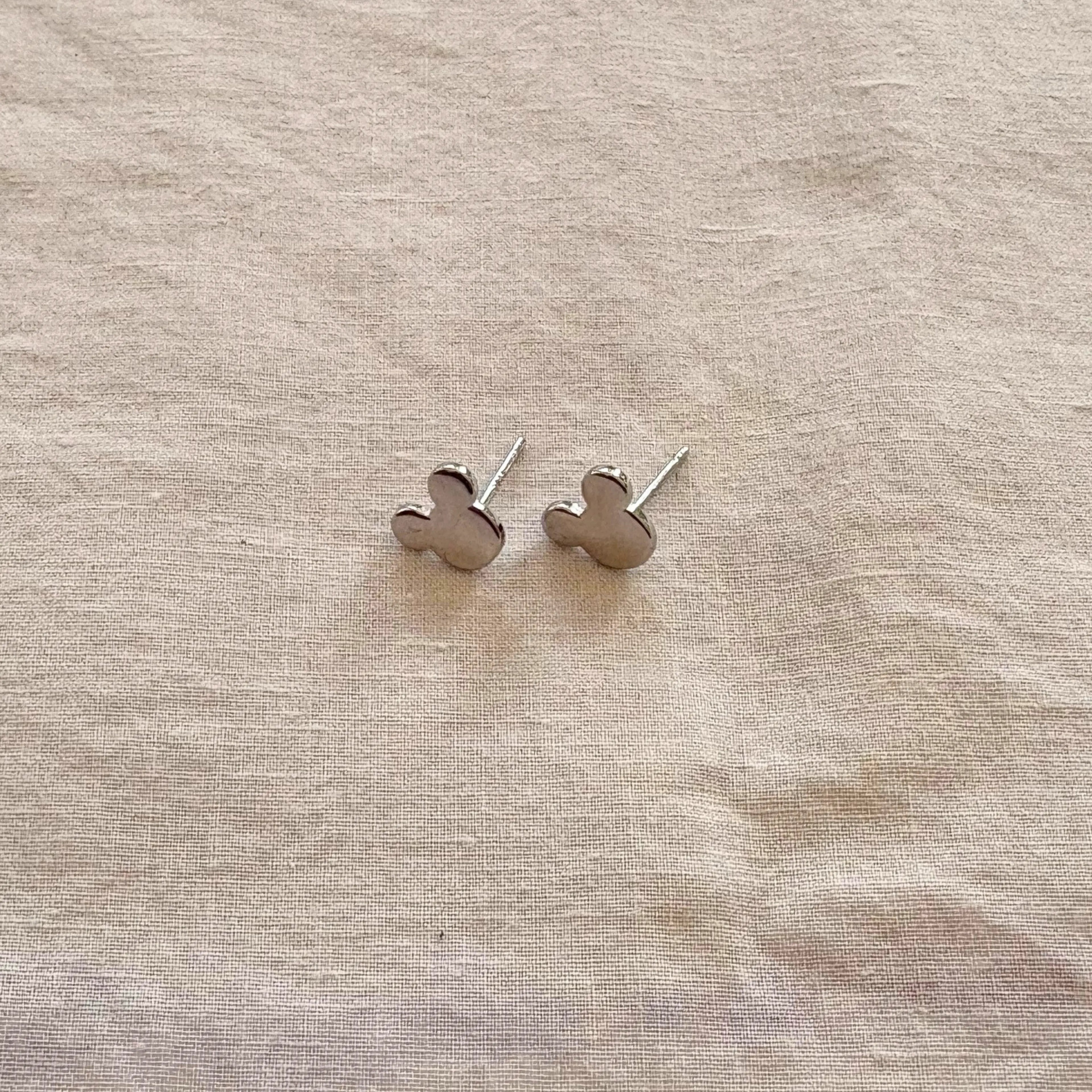 Silver mickey mouse earrings on a beige fabric background