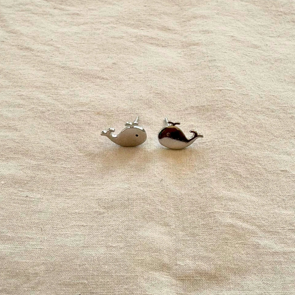 Two small whale-shaped earrings on a beige fabric background