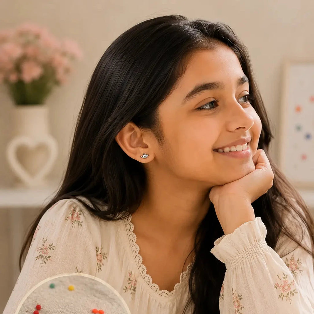 A young girl with long dark hair smiles while resting her chin on her hand, wearing a cream floral blouse. A circular inset at the bottom left shows a pair of small black and silver crescent moon stud earrings on a soft gray fabric background.