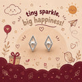 A pair of silver geometric stud earrings with clear crystal-like centers, displayed on a beige circular stand against a whimsical illustrated background with the text 'tiny sparkle, big happiness!'. The background features cartoon clouds, a sun, hearts, stars, a bird, and a gift box.