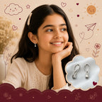 A smiling young girl with dark hair and a pearl hair clip is shown wearing a small silver hoop earring with a clear teardrop-shaped stone. A cloud-shaped inset on the right shows a pair of the earrings on a soft blue-gray fabric.