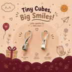 A pair of tiny sterling silver cube-shaped dangle earrings displayed on a decorative beige and burgundy card. The card features cute cartoon illustrations of a sun, cloud, bird, gift box, and stars, along with text that reads 'Tiny Cubes, Big Smiles!' and '92.