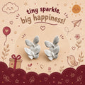 A pair of silver butterfly-shaped stud earrings displayed on a decorative illustrated backing card that reads 'tiny sparkle, big happiness!' with doodles of a sun, cloud, balloon, gift, bird, and stars.