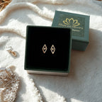 A pair of 925 silver diamond-shaped stud earrings displayed in an open black jewelry box, with the branded green lid placed behind it on a soft white textured blanket.