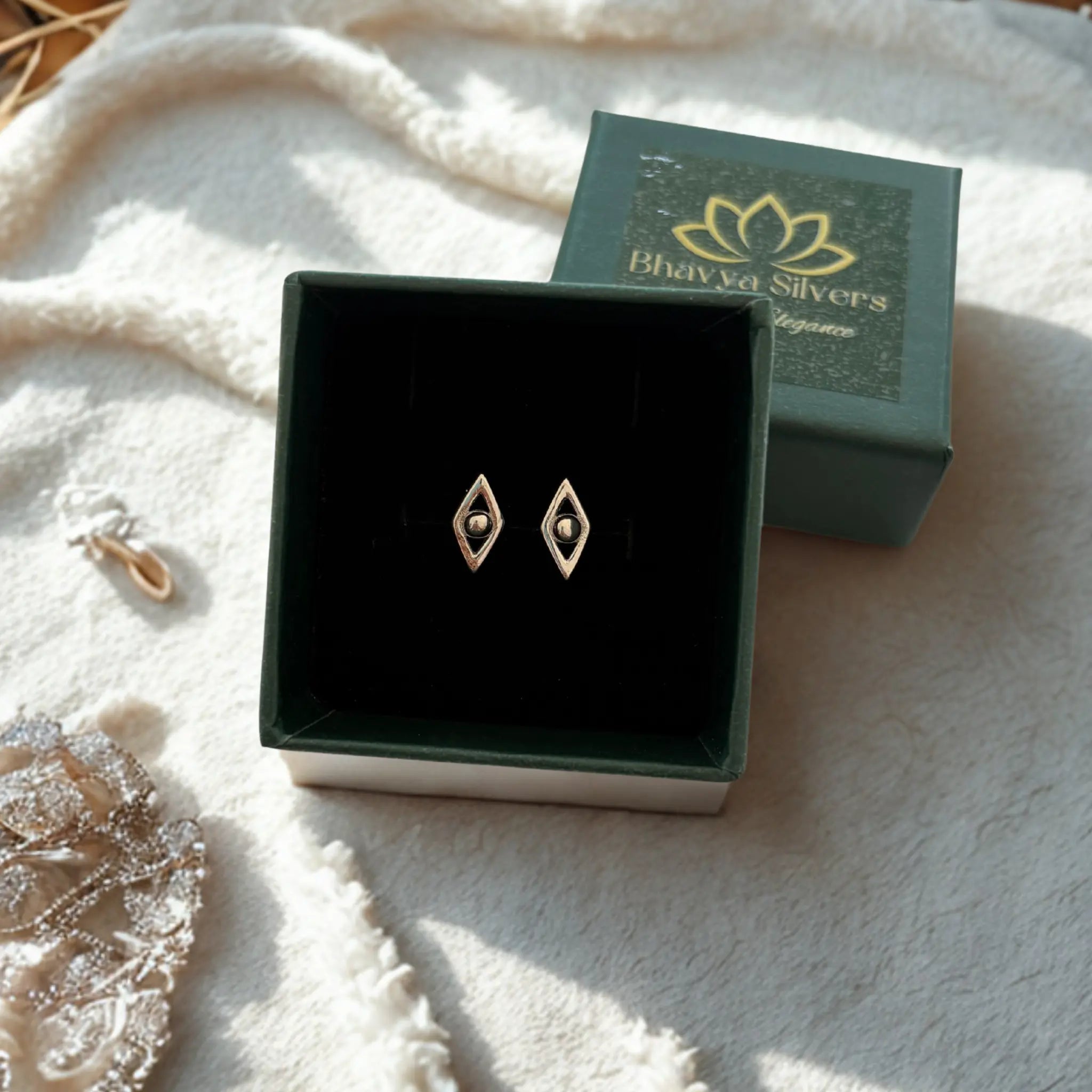 A pair of 925 silver diamond-shaped stud earrings displayed in an open black jewelry box, with the branded green lid placed behind it on a soft white textured blanket.