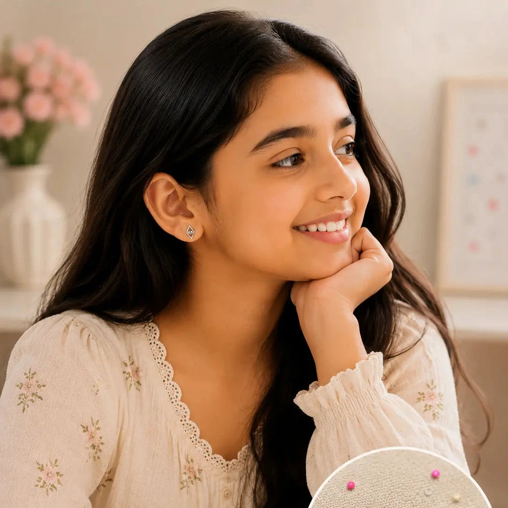 A young girl with long dark hair is smiling and looking to the side, wearing a cream floral blouse. In the lower right, an inset circle shows a pair of silver geometric stud earrings with small colorful bead details pinned onto fabric.