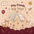 A cute illustrated product graphic showing two small white deer centered on a warm beige background with the text 'tiny friends, big love!' above them. The scene includes cartoon animals, flowers, stars, hearts, a sun, clouds, and decorative border elements, suggesting a jewelry or accessory listing with a whimsical design.