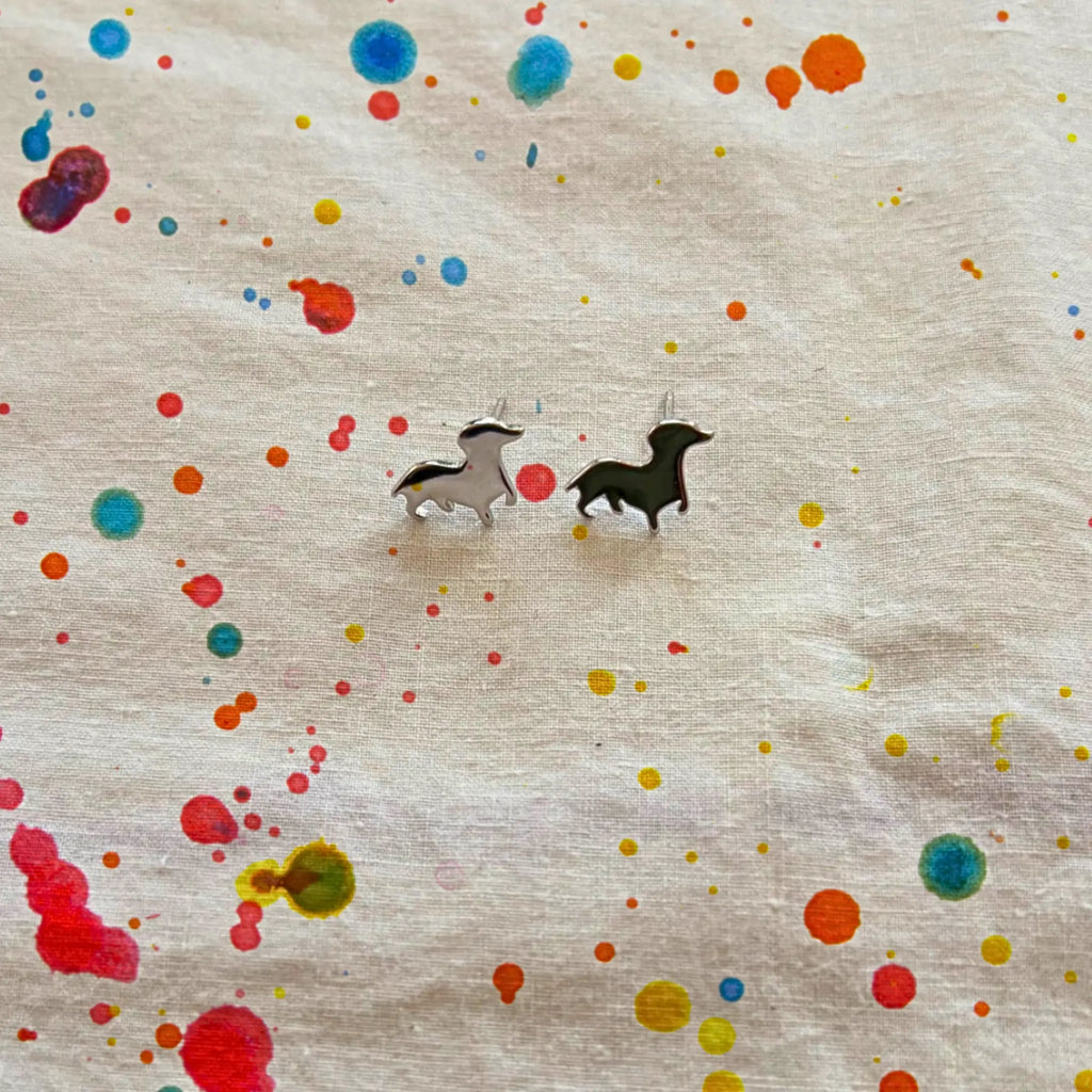 A pair of small animal stud earrings, displayed on a textured cream fabric with colorful paint splatters.