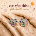 A pair of silver petal-shaped stud earrings with small multicolored gemstones, displayed on a soft beige textured blanket with decorative text and doodles reading 'everyday shine for little ones'.