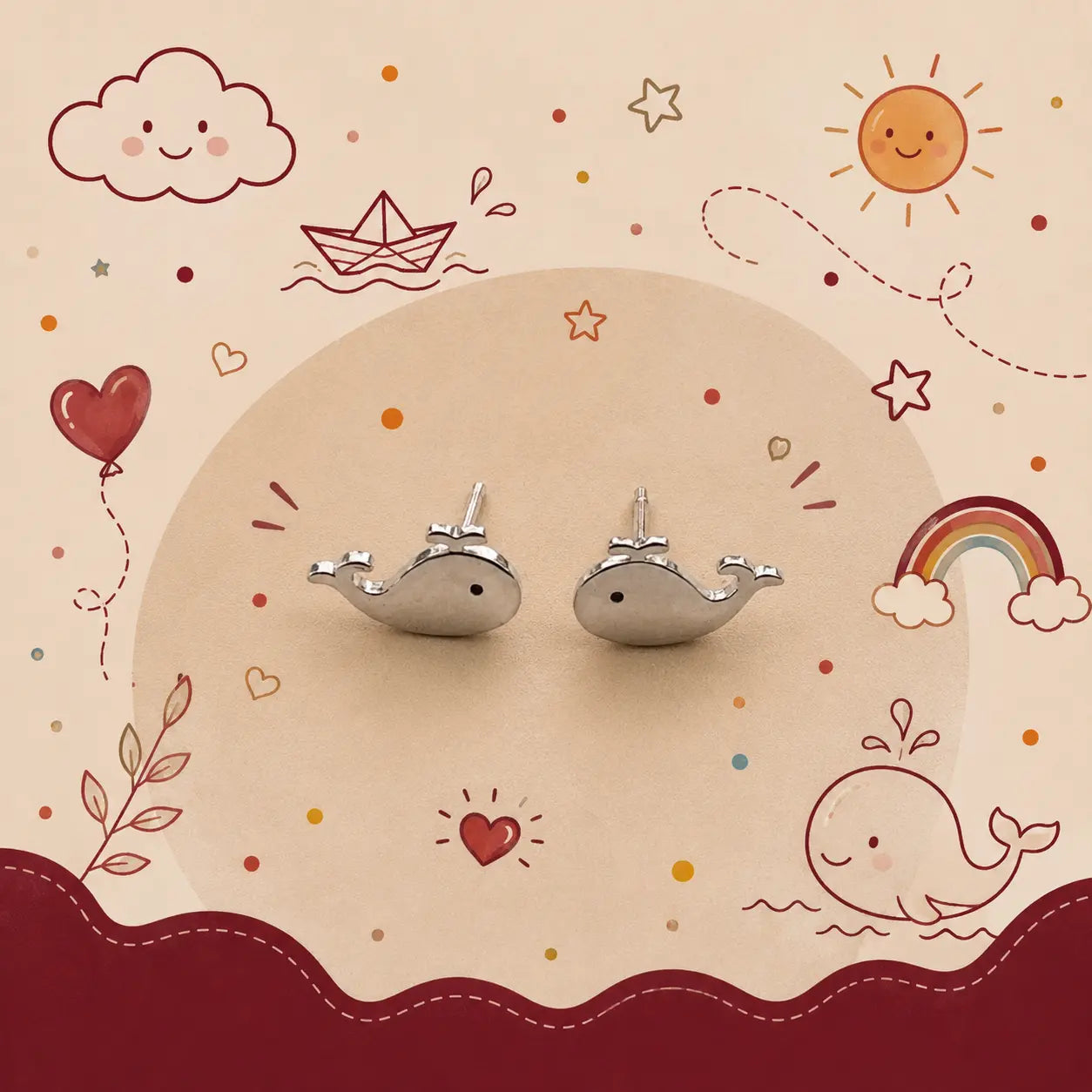 A pair of silver whale-shaped stud earrings displayed on a beige circular card against a whimsical cream background decorated with cartoon clouds, stars, a sun, a rainbow, and a whale illustration.