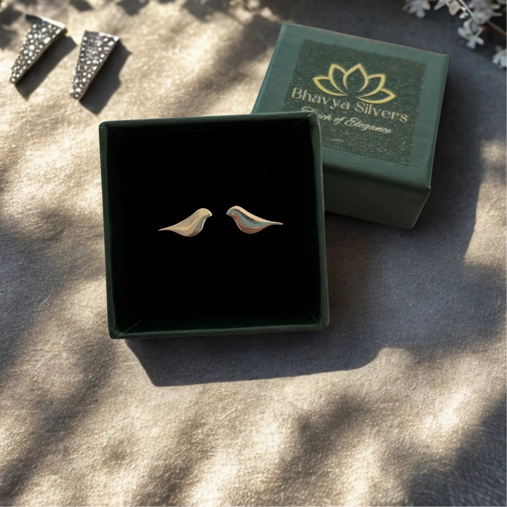 A pair of small bird-shaped silver earrings displayed in an open dark green jewelry box, with the branded box lid placed behind them on a textured beige surface.