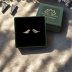 A pair of small bird-shaped silver earrings displayed in an open dark green jewelry box, with the branded box lid placed behind them on a textured beige surface.