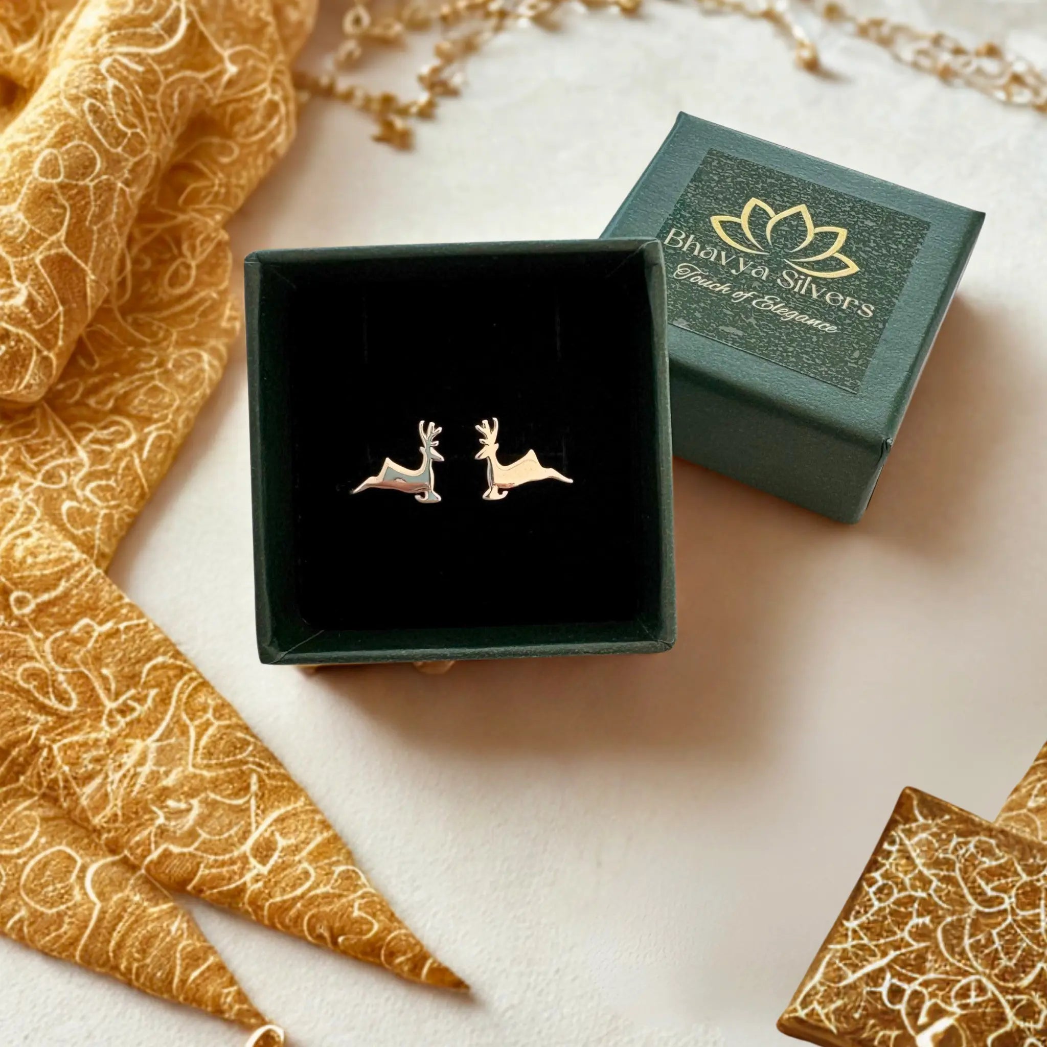 A pair of small deer-shaped stud earrings in silver-tone metal, displayed inside an open dark green jewelry box next to its branded lid. The box is styled on a light beige surface with gold patterned fabric and jewelry props.
