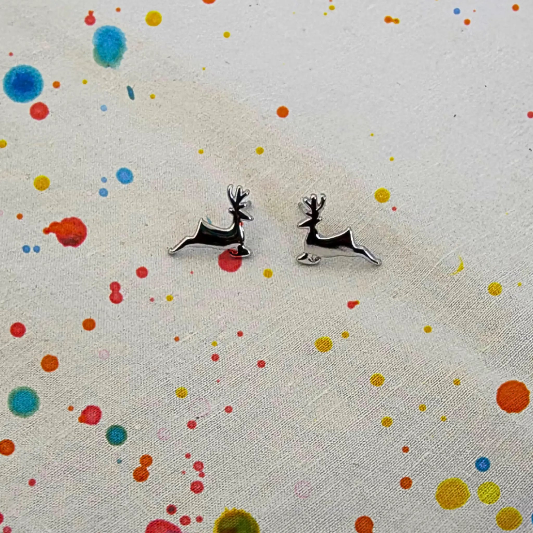 A pair of small silver deer stud earrings with black enamel detailing, displayed on a textured off-white fabric with colorful paint splatters.
