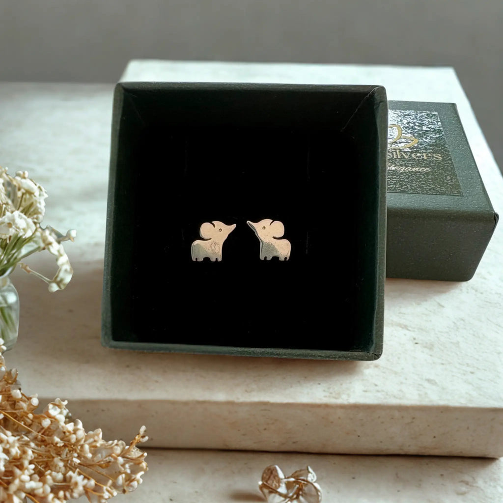 A pair of small elephant-shaped stud earrings in a pale pinkish-gold tone, displayed inside an open black jewelry box on a light stone surface with dried flowers nearby.