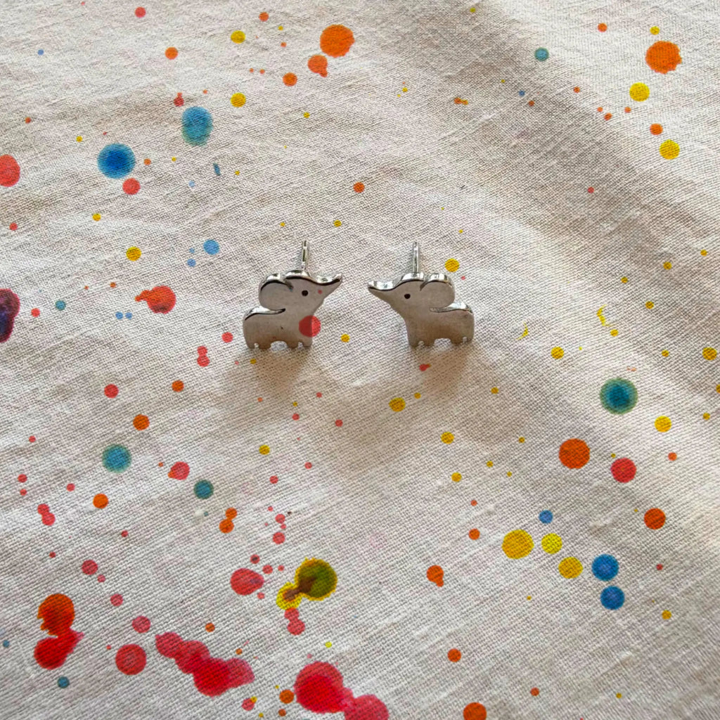 A pair of small silver elephant stud earrings resting on a textured off-white fabric background decorated with colorful paint splatters.