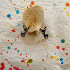 A pair of small black-and-white dog earrings displayed on a beige fabric with colorful paint splatters, next to a natural seashell.