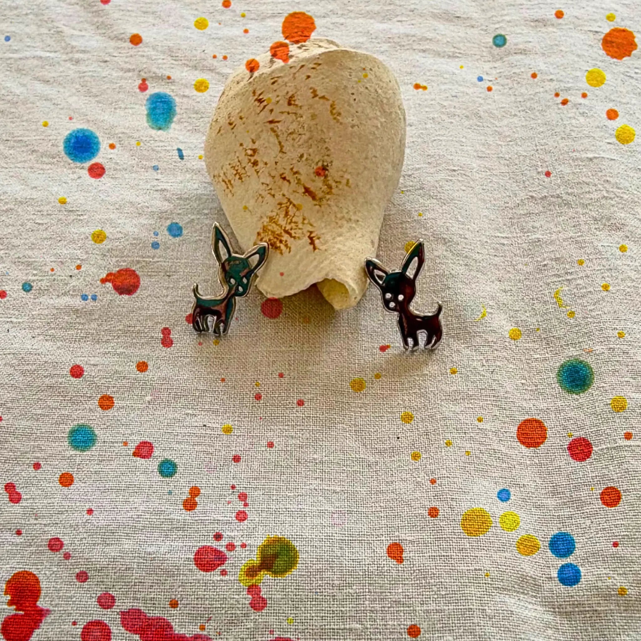 A pair of small black-and-white dog earrings displayed on a beige fabric with colorful paint splatters, next to a natural seashell.