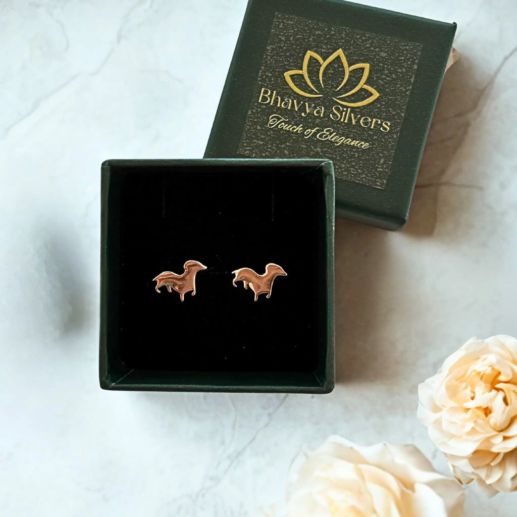 A pair of small silver puppy-shaped stud earrings displayed in an open green jewelry box, with the branded dark green lid placed behind it on a light marble surface. Decorative cream flowers appear in the lower right corner.