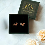 A pair of small silver puppy-shaped stud earrings displayed in an open green jewelry box, with the branded dark green lid placed behind it on a light marble surface. Decorative cream flowers appear in the lower right corner.