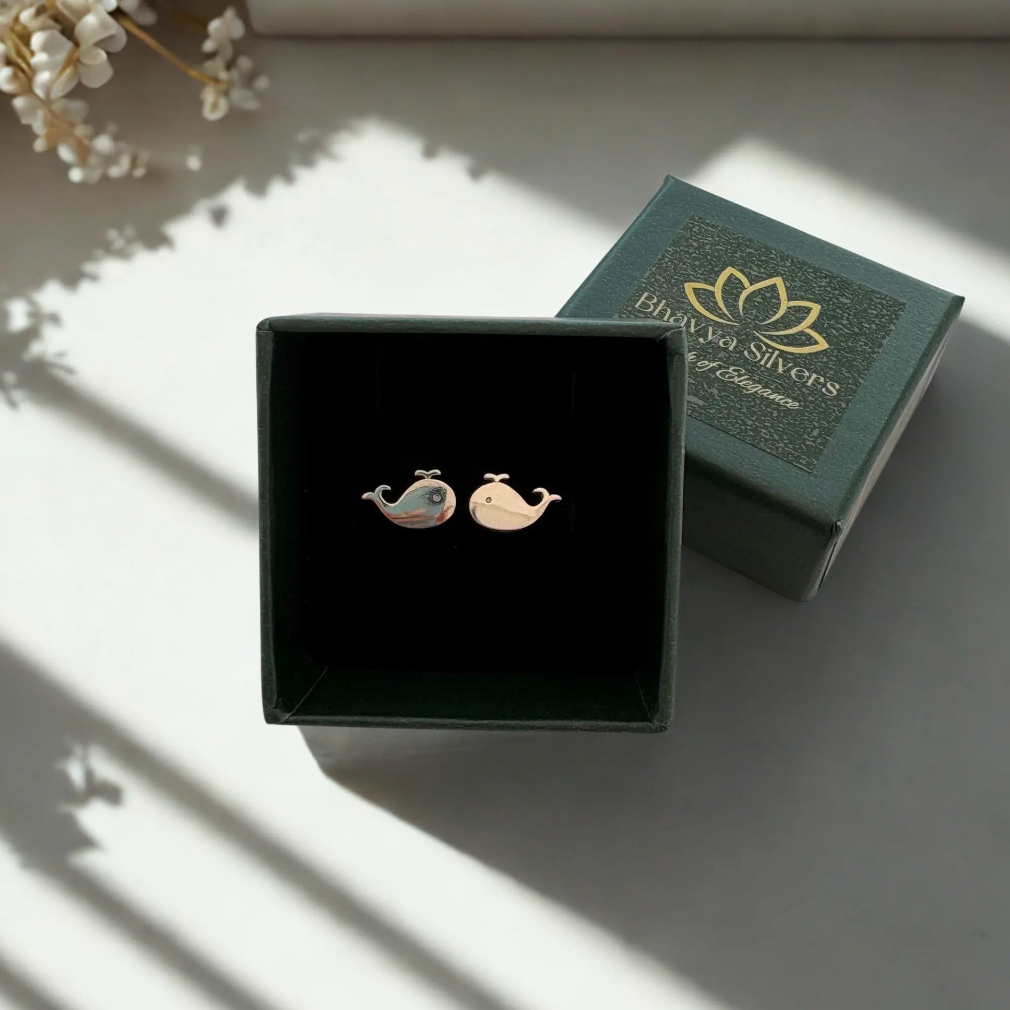 A pair of whale-shaped stud earrings displayed in an open dark green jewelry box, with the branded box lid placed behind it on a softly lit surface.
